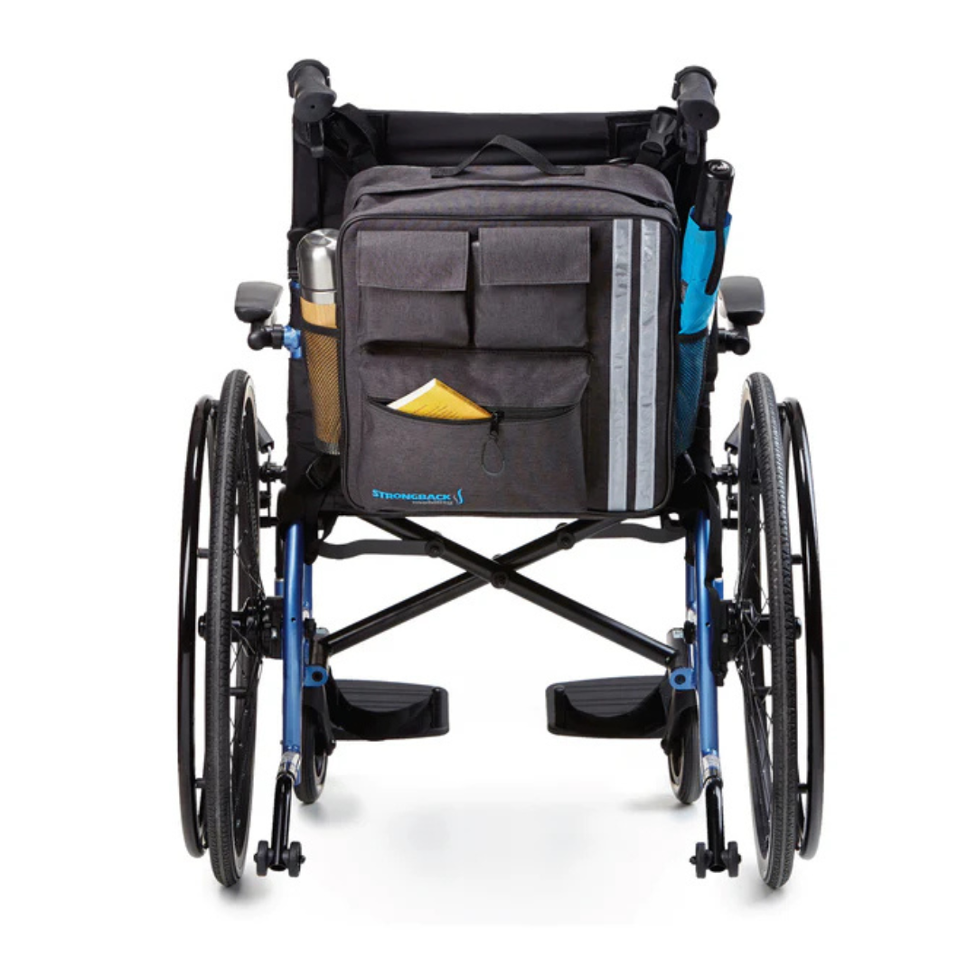 A Strongback Travel Storage Bag For Mobility Aids is attached to the wheelchair backrest, featuring multiple pockets and holding a water bottle, an umbrella, and a yellow item in the front pocket.